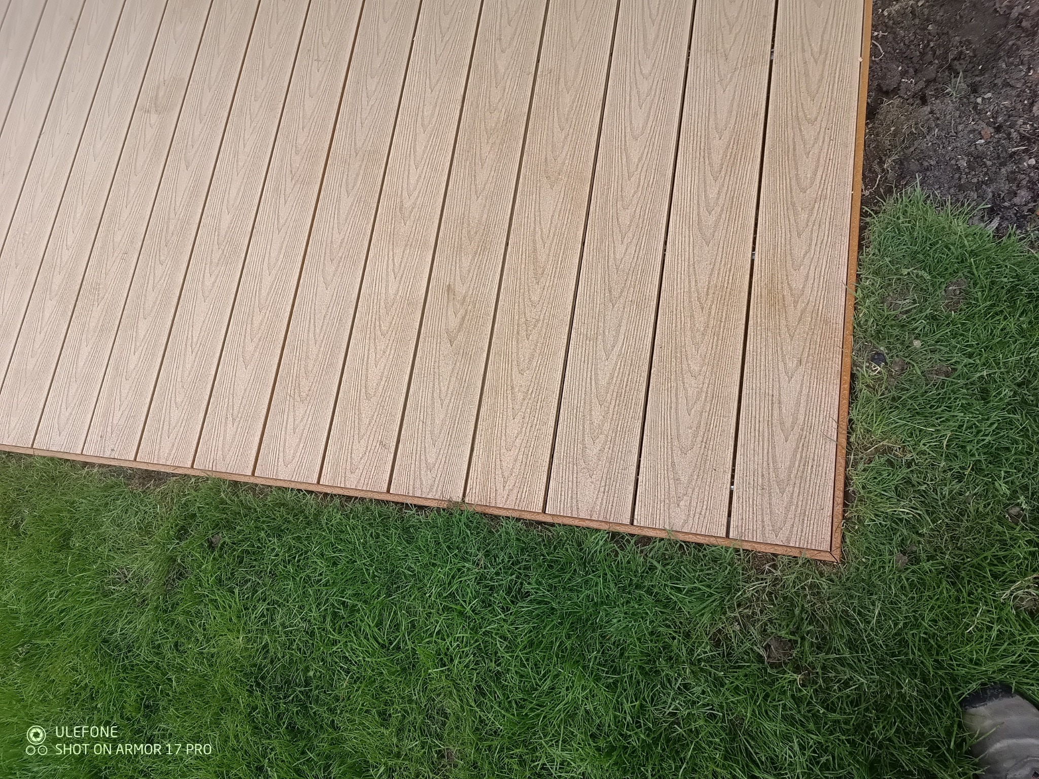 Jasny taras kompozytowy z bliska, z widoczną strukturą desek i zieloną trawą obok, świeżo po montażu, z minimalnym widokiem ziemi.