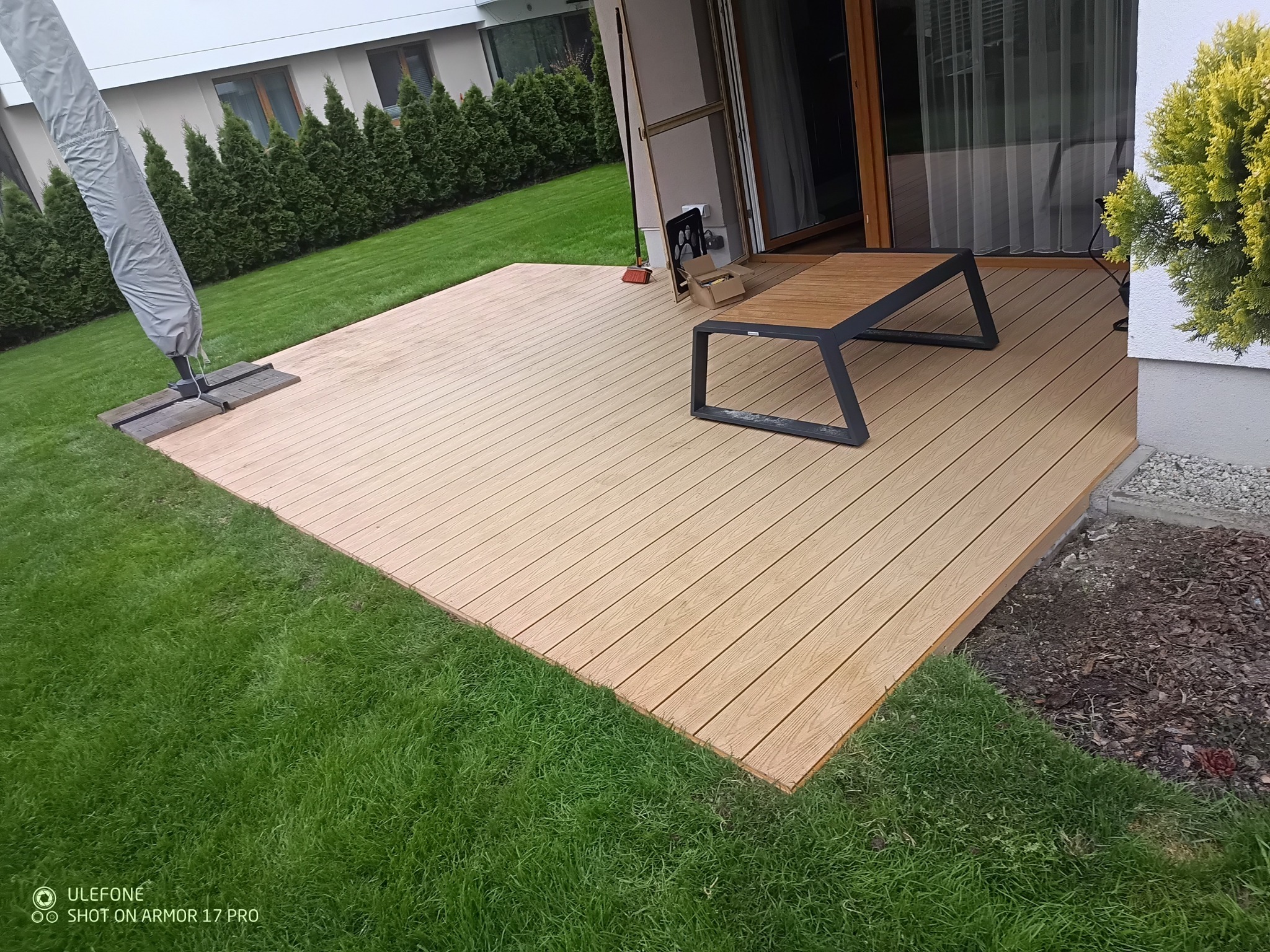 Jasny taras kompozytowy z nowoczesnym stolikiem na tle zielonego trawnika i domu. Widok z góry, oświetlenie dzienne.