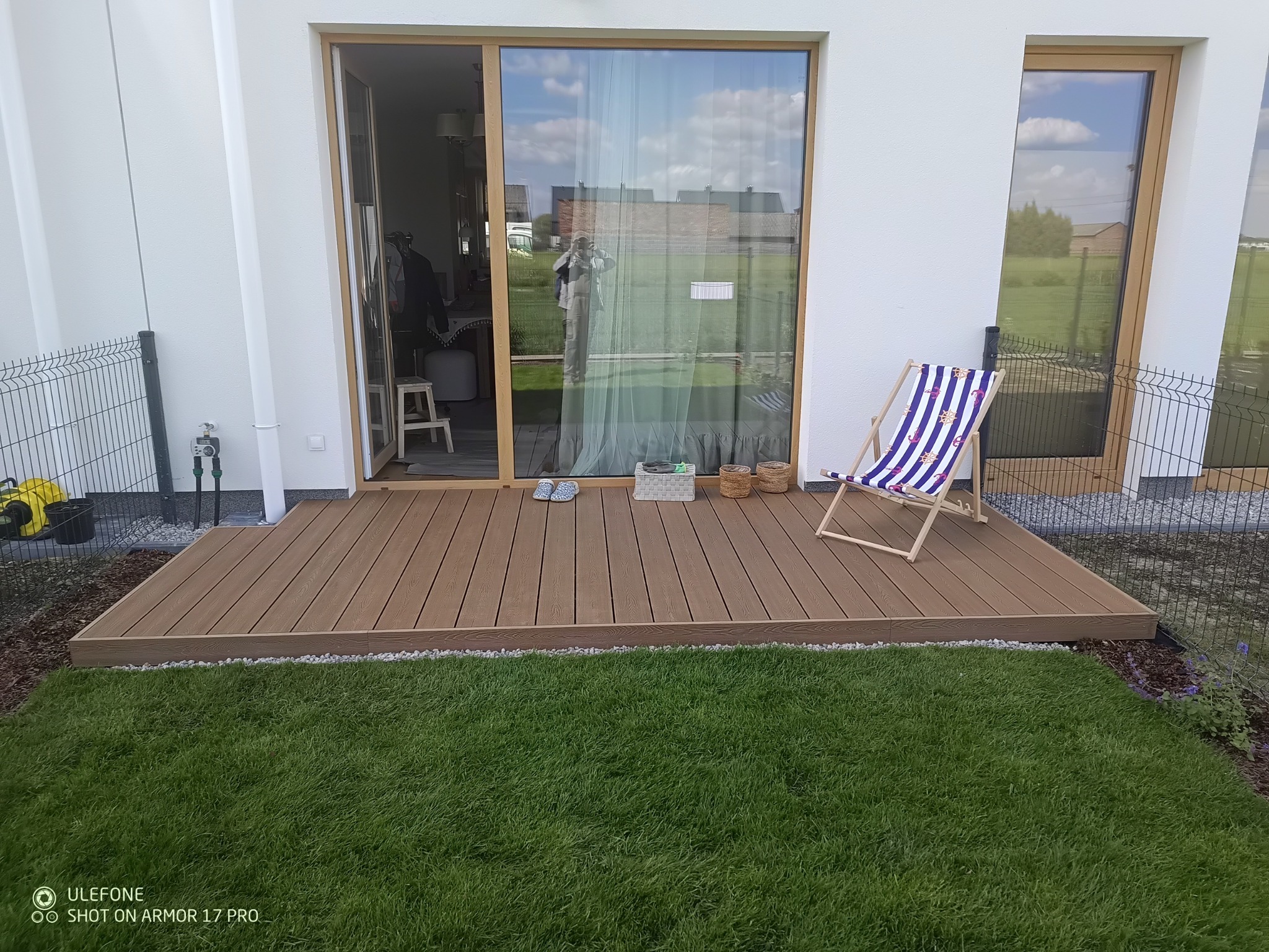 Nowoczesny taras z deski kompozytowej w Warszawie, z leżakiem w paski i widokiem na trawnik. Uwagę przyciągają przesuwne drzwi z refleksami.