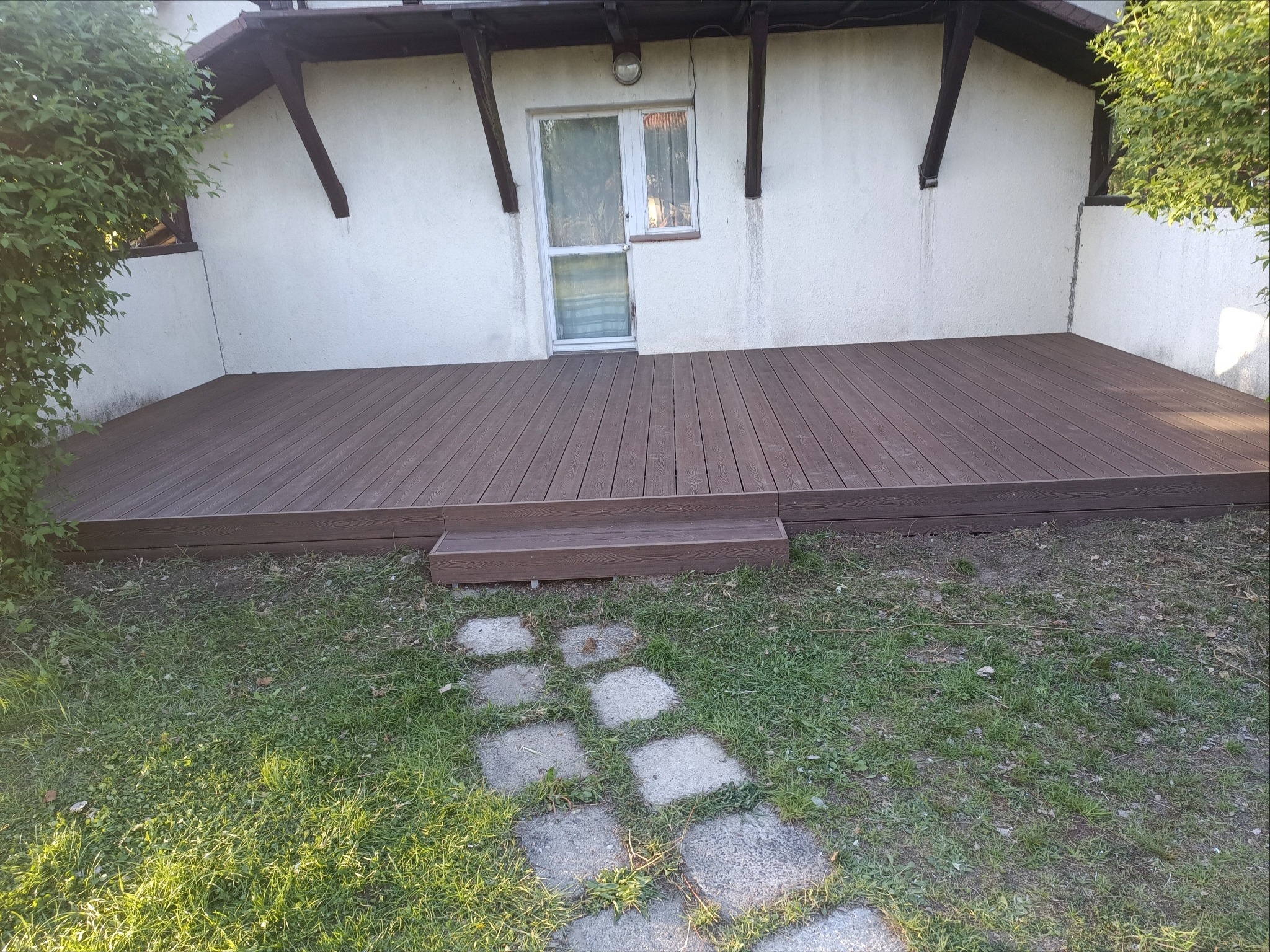 Brunatny taras kompozytowy z pojedynczym stopniem, przylegający do białej ściany budynku z oknem. Kamienna ścieżka prowadzi przez trawnik.