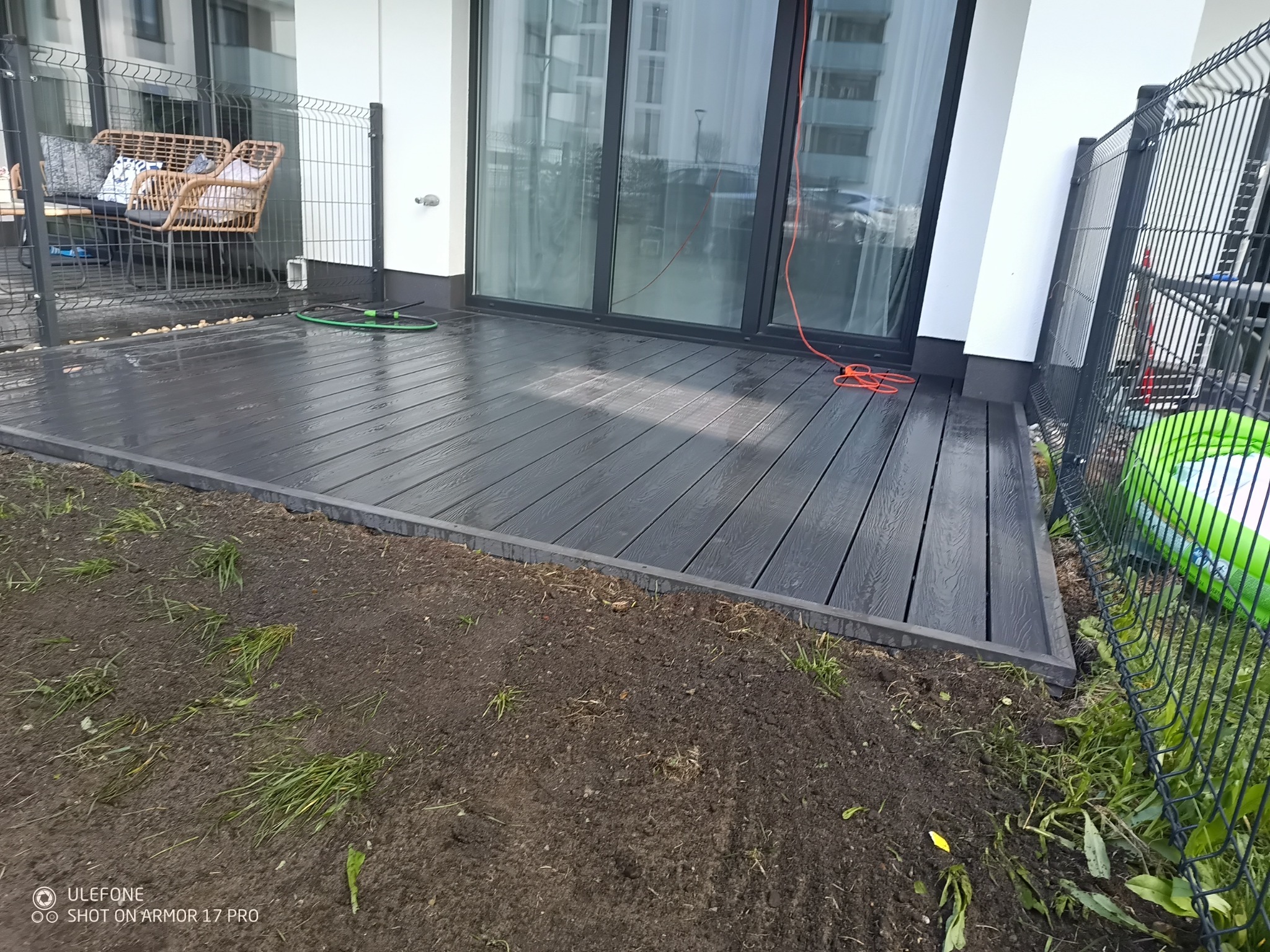 Nowoczesny, grafitowy taras z desek kompozytowych przylegający do budynku z dużymi przeszkleniami. Widoczne meble ogrodowe i trawnik.