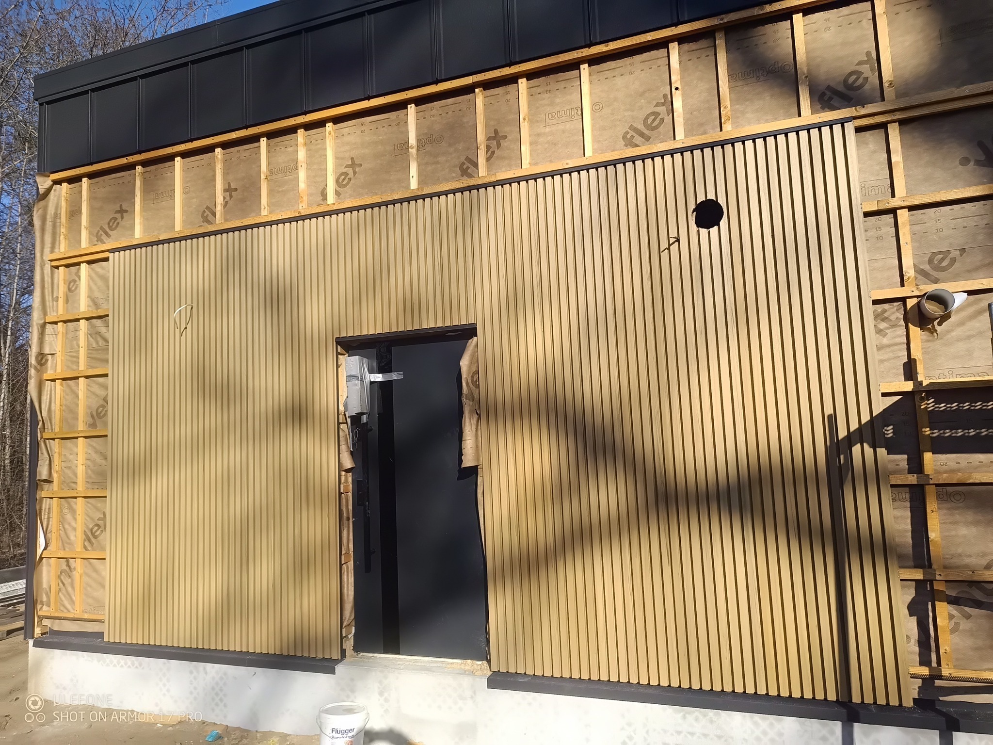 Montaż elewacji z drewnianych listew na budynku. Widoczne elementy konstrukcyjne i izolacja. Częściowo zamontowane listwy tworzą pionowy wzór.