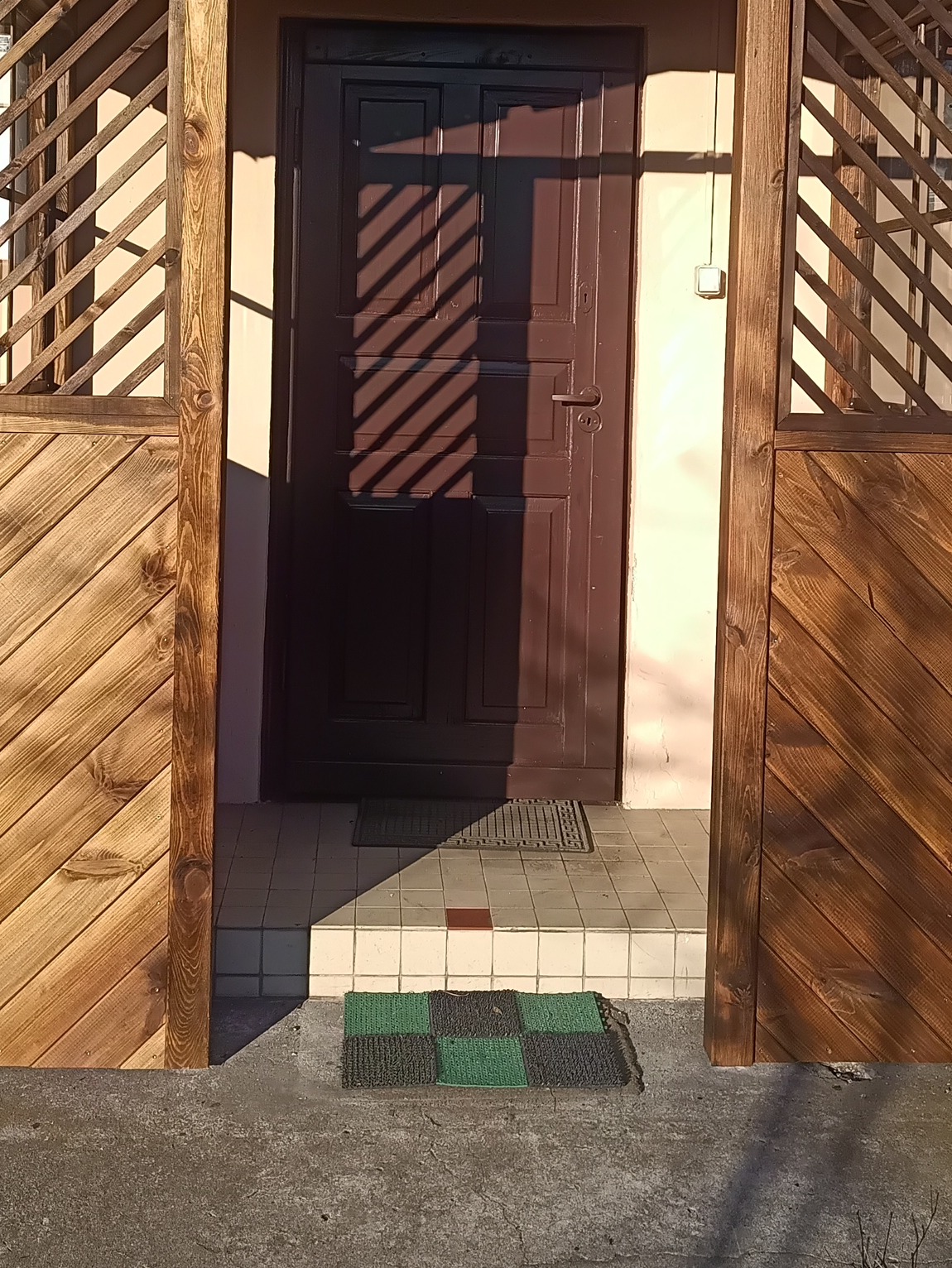 Wejście do budynku z drewnianą zabudową, brązowe drzwi z cieniem, płytki i wycieraczka. Elementy drewniane w ciepłym odcieniu brązu.