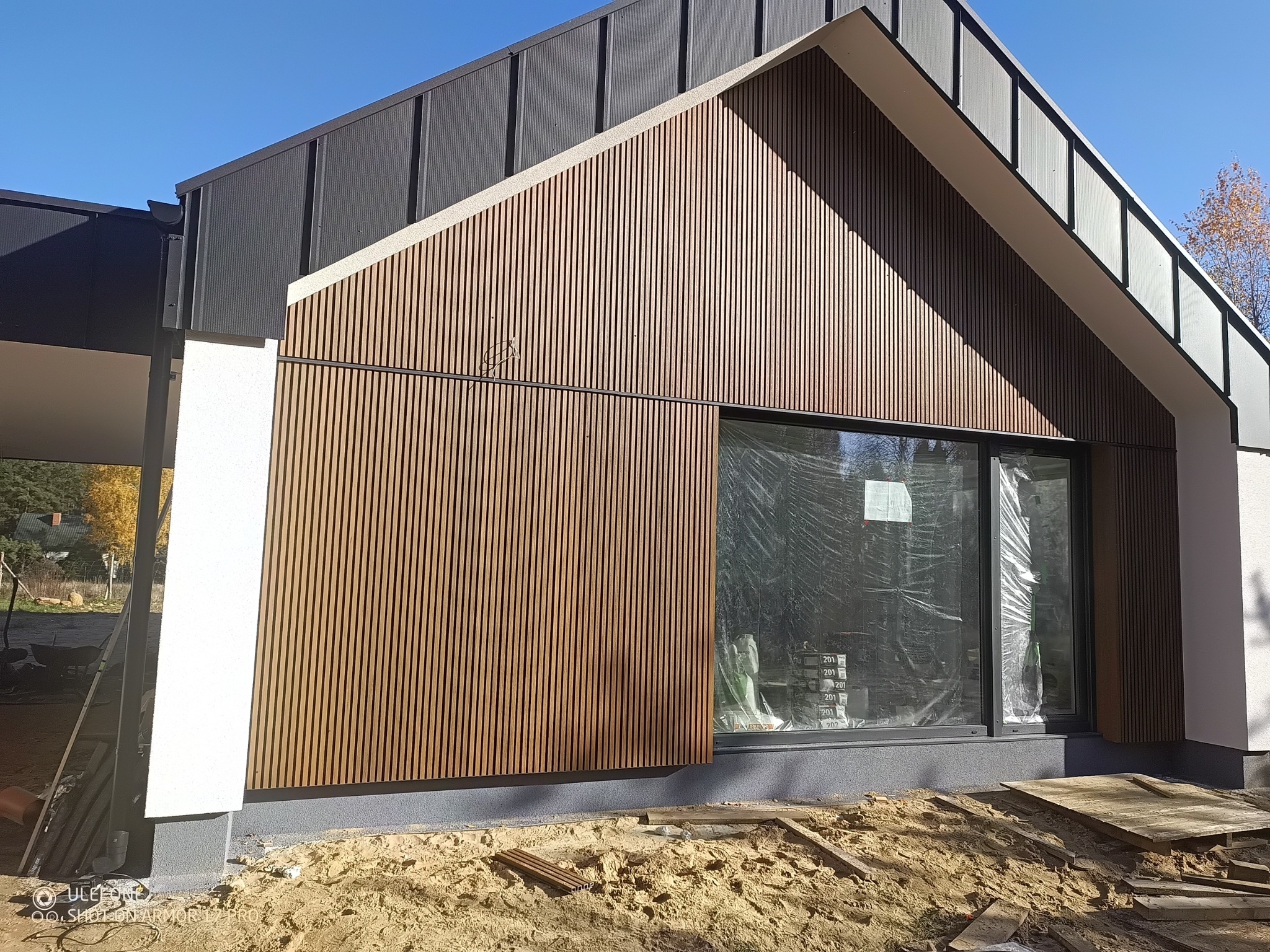 Nowoczesna elewacja domu z drewnianymi panelami pionowymi w ciepłym odcieniu brązu, okno zabezpieczone folią, jasne tynki i grafitowe elementy dachu.