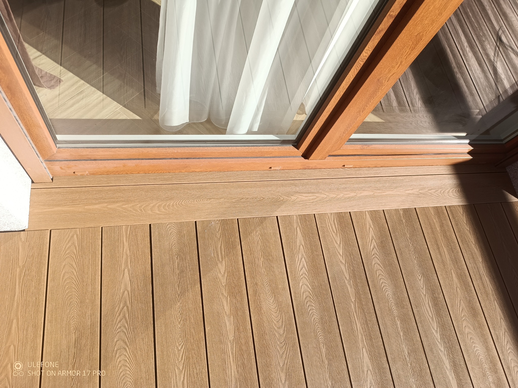 Jasny taras kompozytowy z widocznym fragmentem okna balkonowego i firanką. Deski ułożone równolegle, z delikatnym wzorem imitującym drewno. Słoneczny dzień.