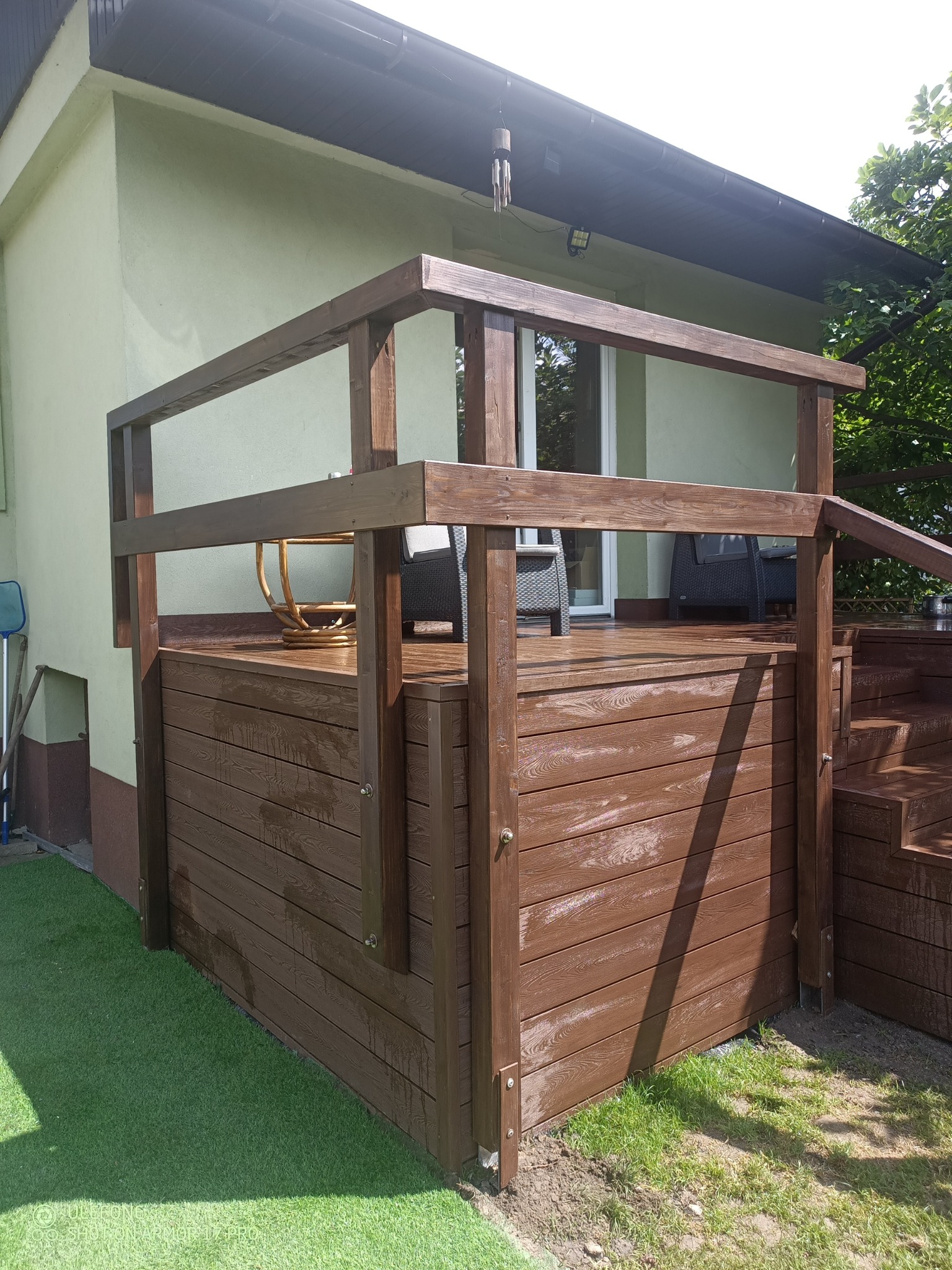 Nowoczesny taras kompozytowy z brązowymi deskami, z niską balustradą i schodami, przylegający do budynku z zieloną elewacją. Widoczne meble ogrodowe.