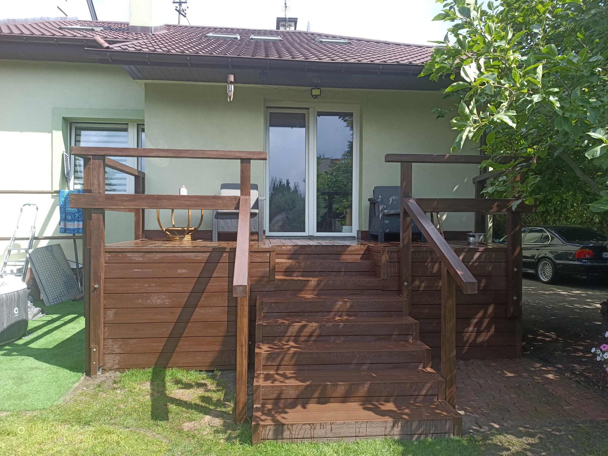 Nowoczesny taras z brązowych desek kompozytowych z schodami, prowadzący do domu z zieloną elewacją. Widoczne meble ogrodowe i fragment samochodu.