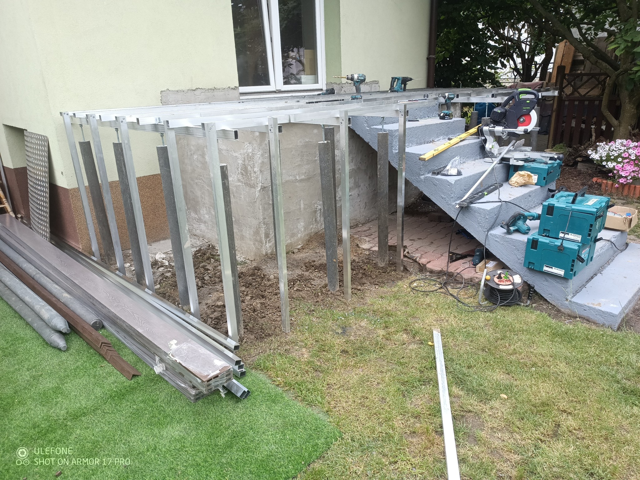Montaż konstrukcji tarasu: aluminiowe profile, deski kompozytowe obok. Widoczne narzędzia na schodach. Etap budowy tarasu przy domu z zielonym trawnikiem.
