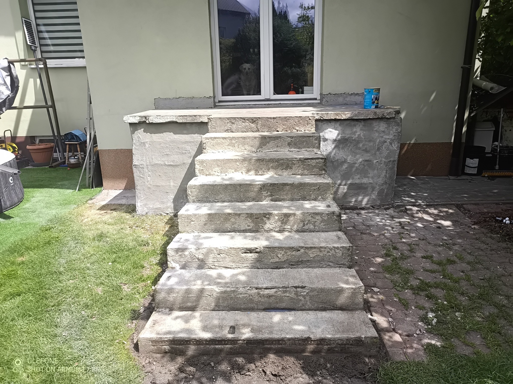 Betonowe schody wejściowe do domu, widoczne ślady napraw i renowacji. Na górze puszka farby, w oknie odbija się pies. Prace remontowe w toku.