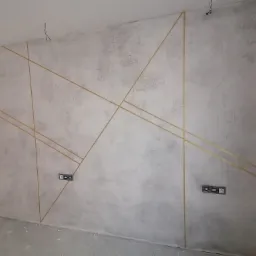 Ściana z szarą gładzią dekoracyjną i złotymi, ukośnymi paskami, widoczne gniazdka elektryczne, surowy stan wykończenia.