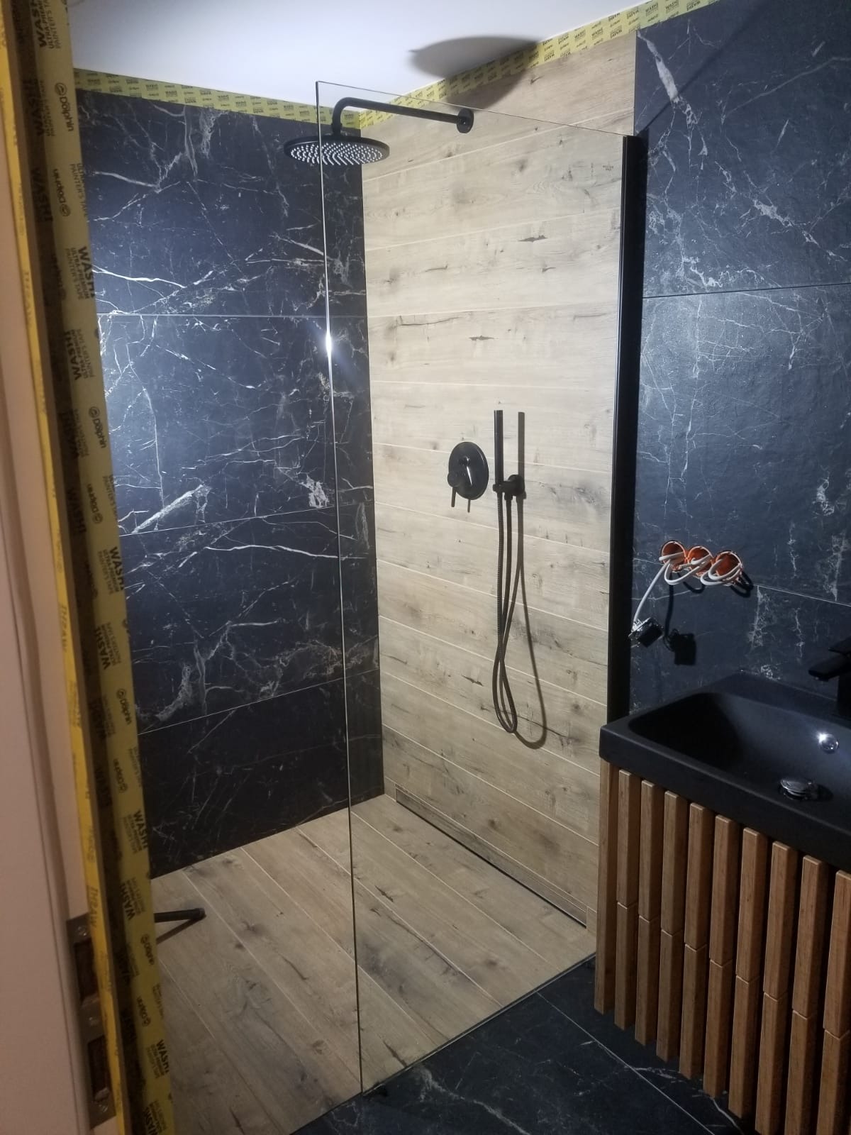 Nowoczesna kabina prysznicowa walk-in z czarną armaturą, obłożona płytkami imitującymi marmur i drewno, z widoczną szafką umywalkową z lamelami.