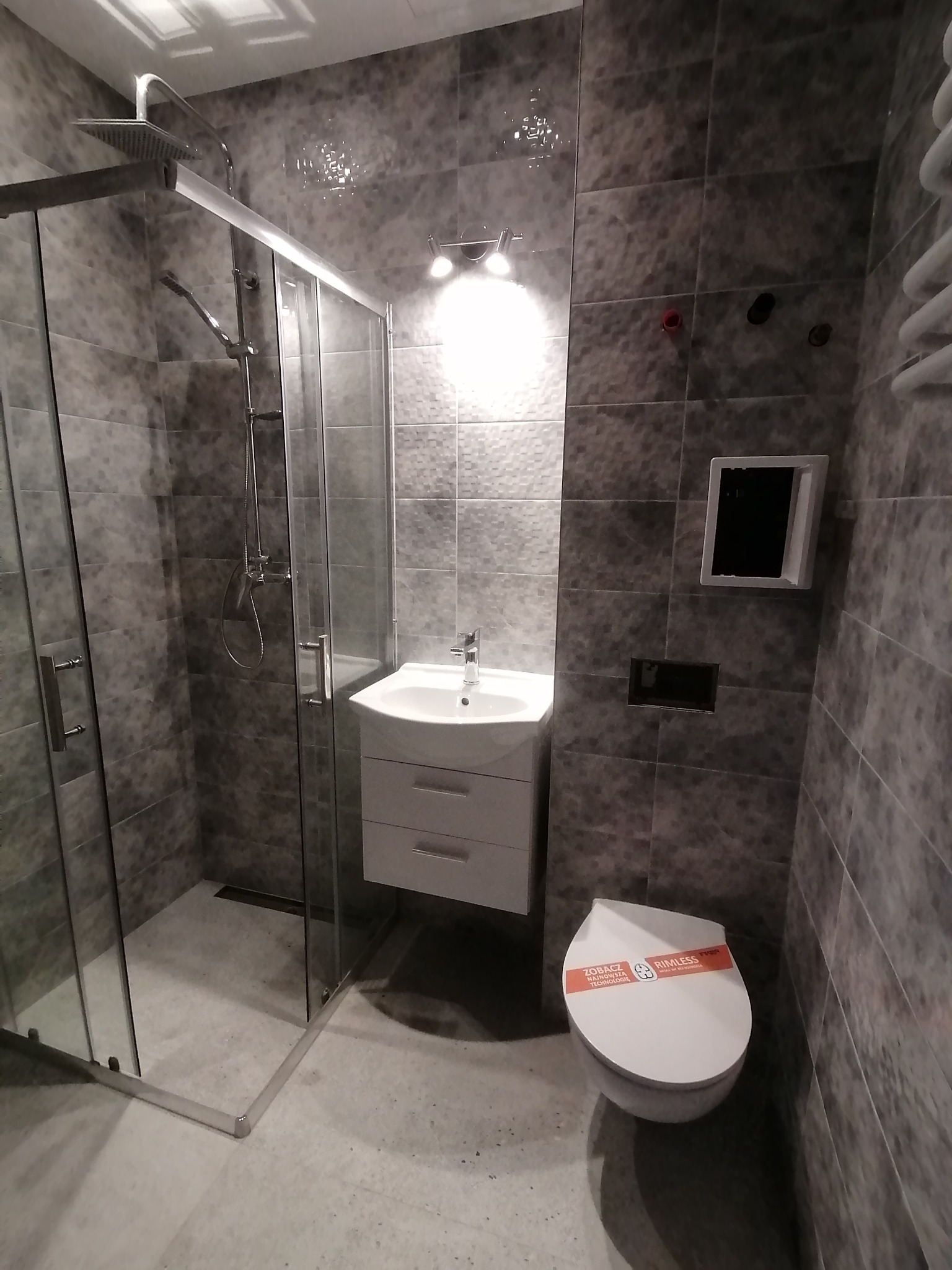 Szara łazienka po remoncie: kabina prysznicowa ze szklanymi drzwiami, umywalka z szafką, wisząca toaleta z naklejką informacyjną, płytki imitujące kamień na ścianach.