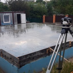 Mazur Vitalii - Świeżo wylana, mokra posadzka zbrojona, otoczona szalunkiem z niebieską izolacją, widoczny niwelator optyczny na statywie, w tle budynek gospodarczy i ogrodzenie z cegły.