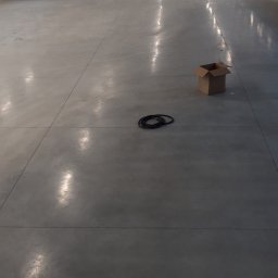 Mazur Vitalii - Duża, pusta hala z szarą posadzką, widoczne ślady po maszynach, zwinięty kabel i kartonowe pudełko na środku, w tle stoły do gry.