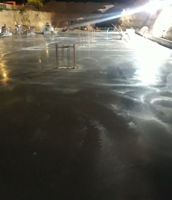 Świeżo wylana, mokra posadzka betonowa w hali przemysłowej, z widocznymi zbrojeniami i maszynami do zacierania w tle, oświetlona sztucznym światłem.