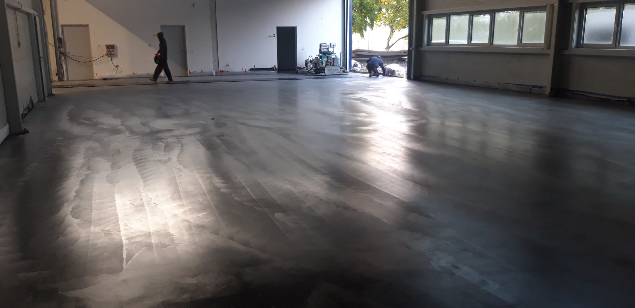 Świeżo wylana, mokra posadzka w hali przemysłowej, z widocznymi śladami po pracy maszyny wygładzającej, dwie osoby w tle oraz maszyna do obróbki posadzek.