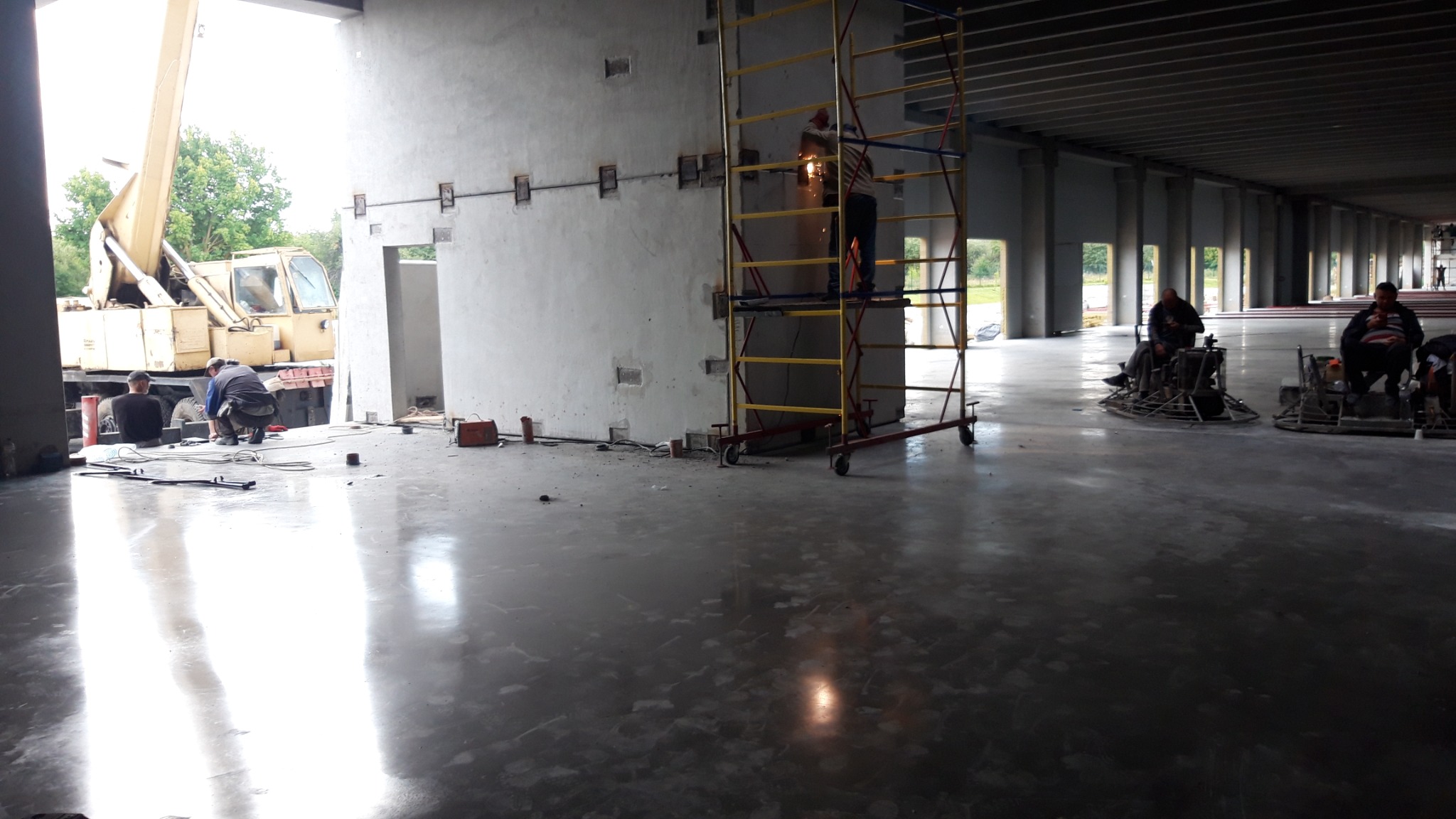 Prace wykończeniowe w hali: spawanie konstrukcji stalowej na rusztowaniu, szlifowanie posadzki jastrychowej za pomocą zacieraczek mechanicznych, widoczny dźwig i pracownicy.