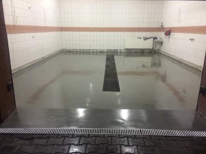 Gładka, szara posadzka z odpływem liniowym i kratką ściekową w pomieszczeniu wyłożonym białymi płytkami z brązowym pasem dekoracyjnym.