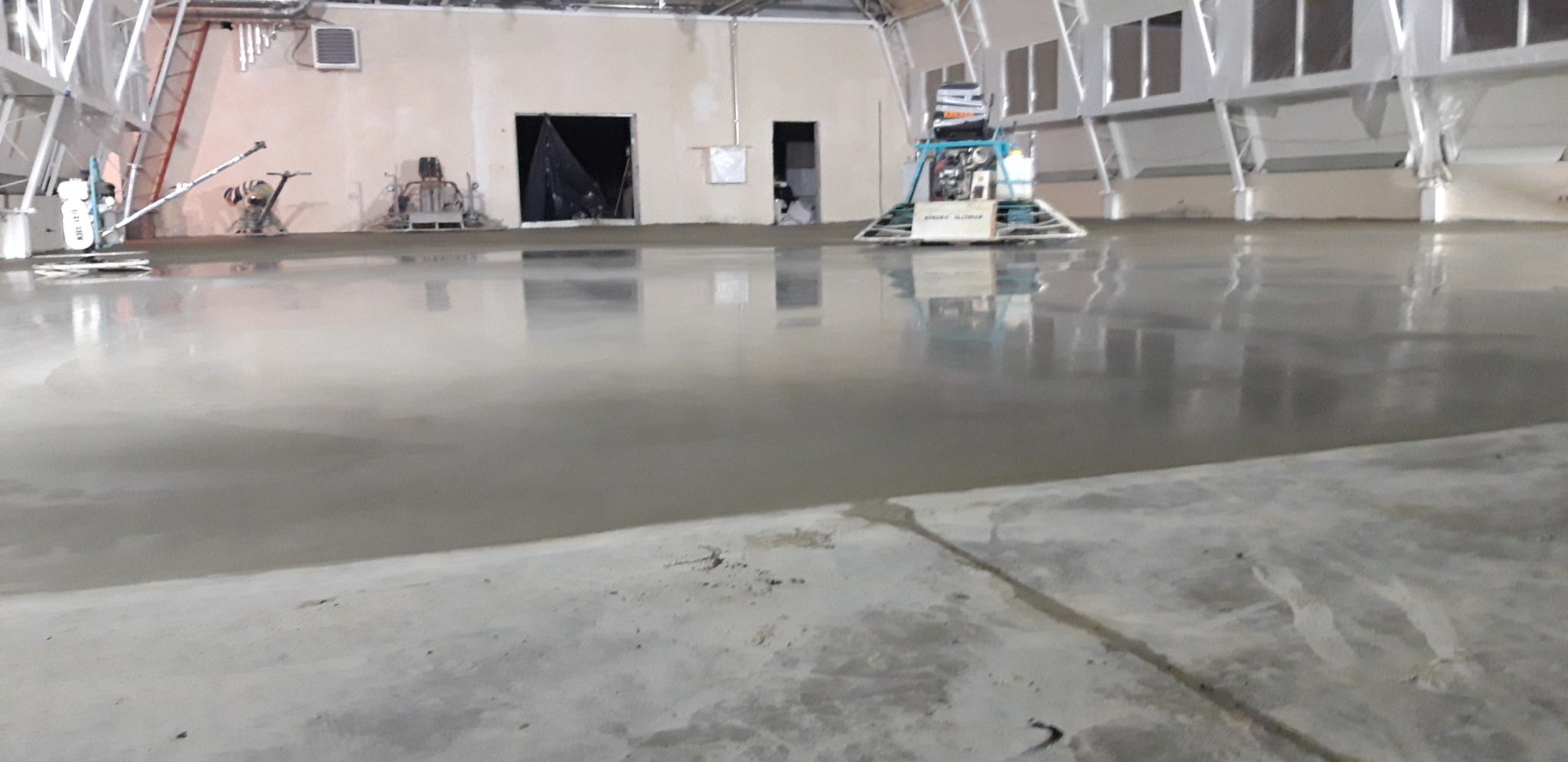 Świeżo wylana, mokra posadzka jastrychowa w hali przemysłowej, maszyna do zacierania w tle, widoczne odbicia w mokrej powierzchni.