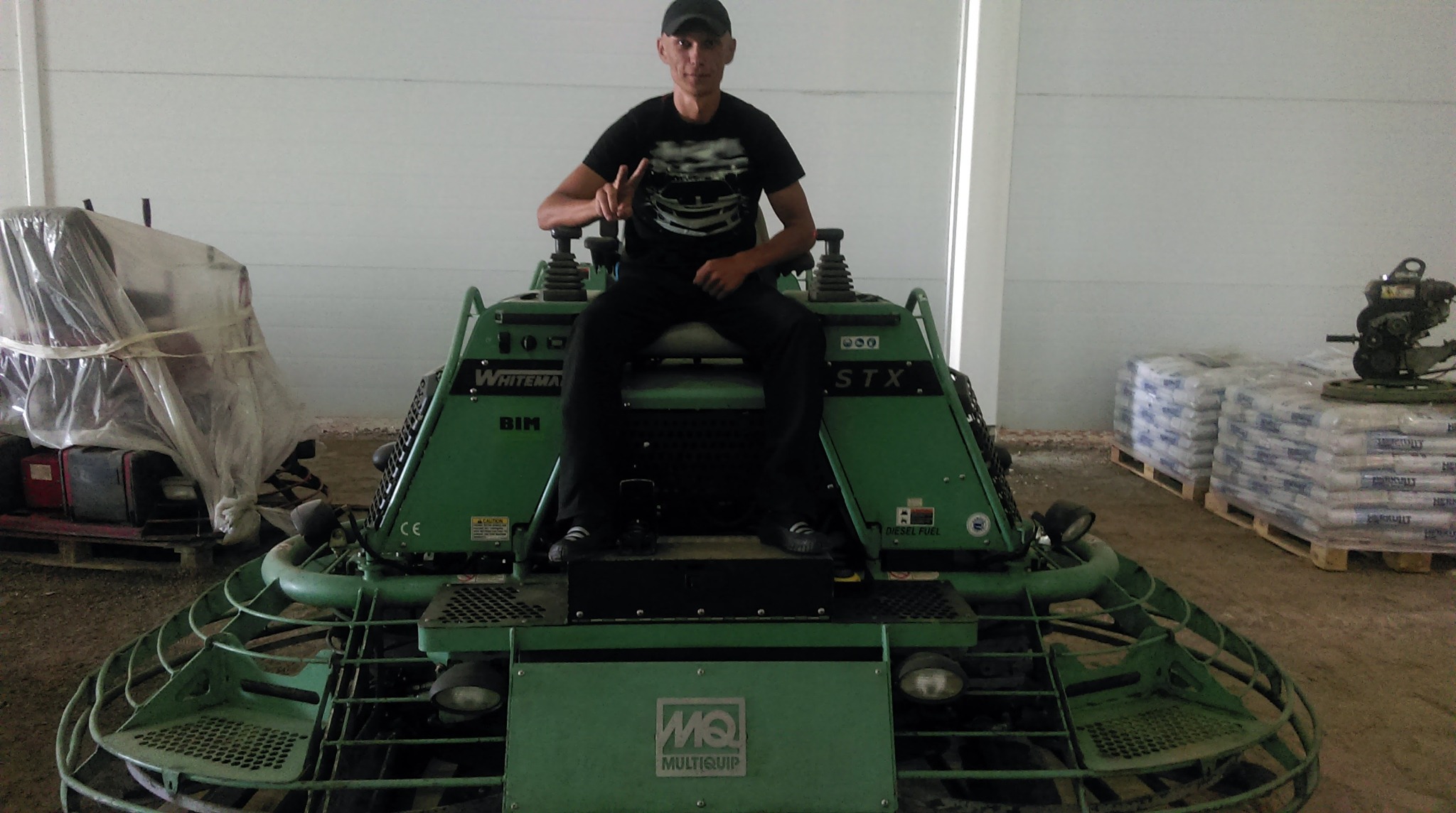 Operator siedzi na zielonej zacieraczce do betonu Whiteman STX BIM, w tle worki z materiałem budowlanym i przykryta folią maszyna.