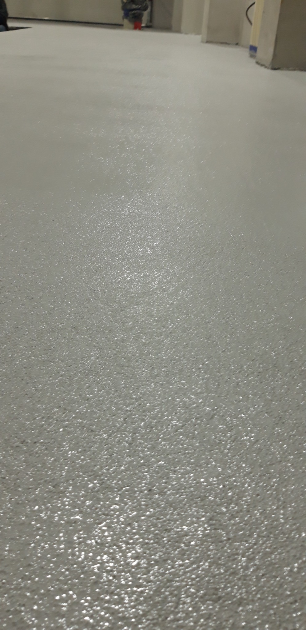 Świeżo wylana, jasnoszara posadzka z widoczną strukturą, odbijająca światło, z fragmentem ściany i słupa w tle.