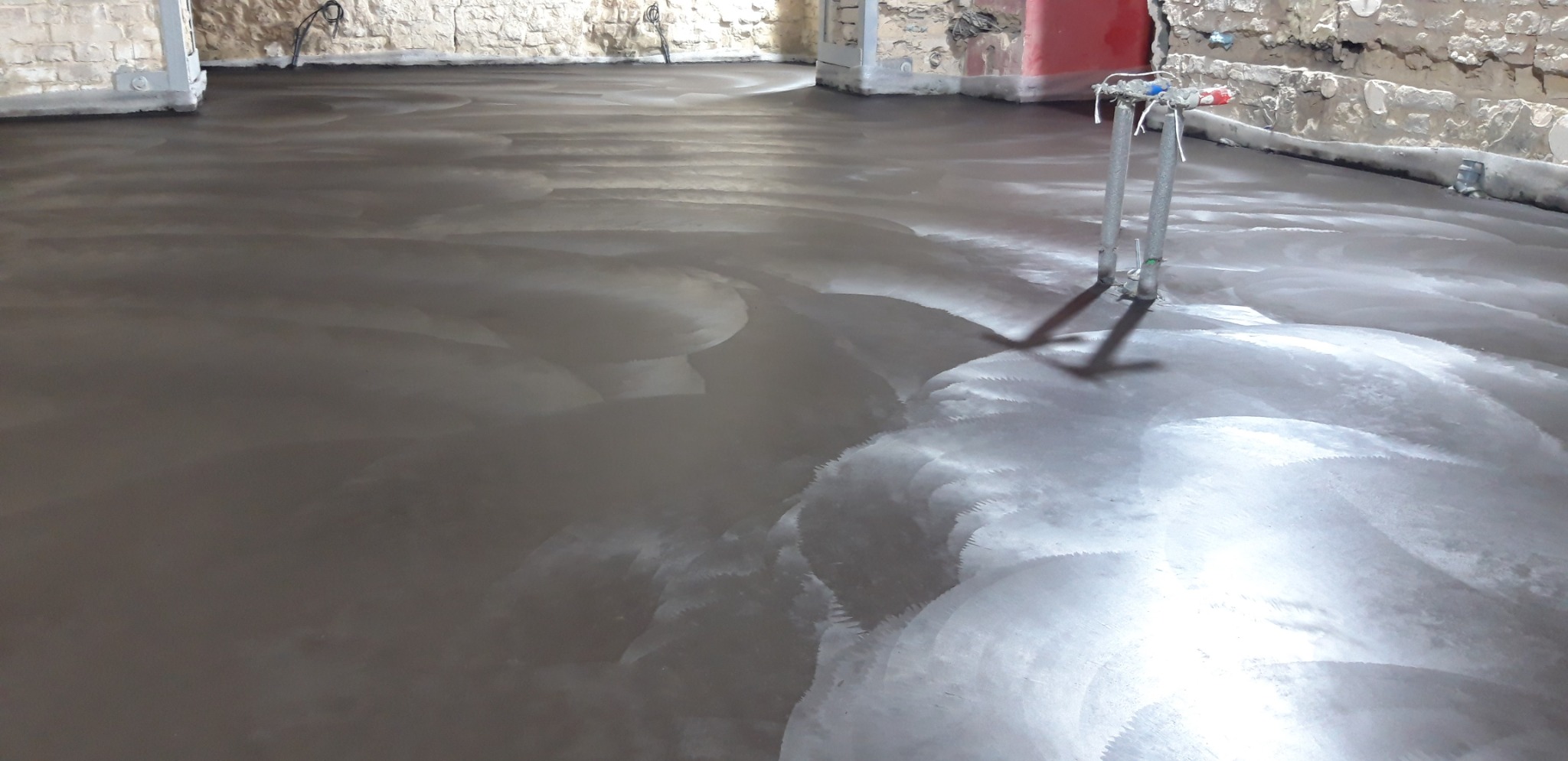 Świeżo wylana, mokra posadzka w surowym wnętrzu z odsłoniętą cegłą i wystającymi rurami, widoczne ślady po rozprowadzaniu materiału.