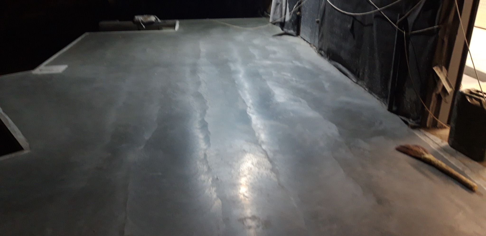 Świeżo wylana, szara posadzka z widocznymi smugami i odbiciami światła, przy ścianie leży miotła.