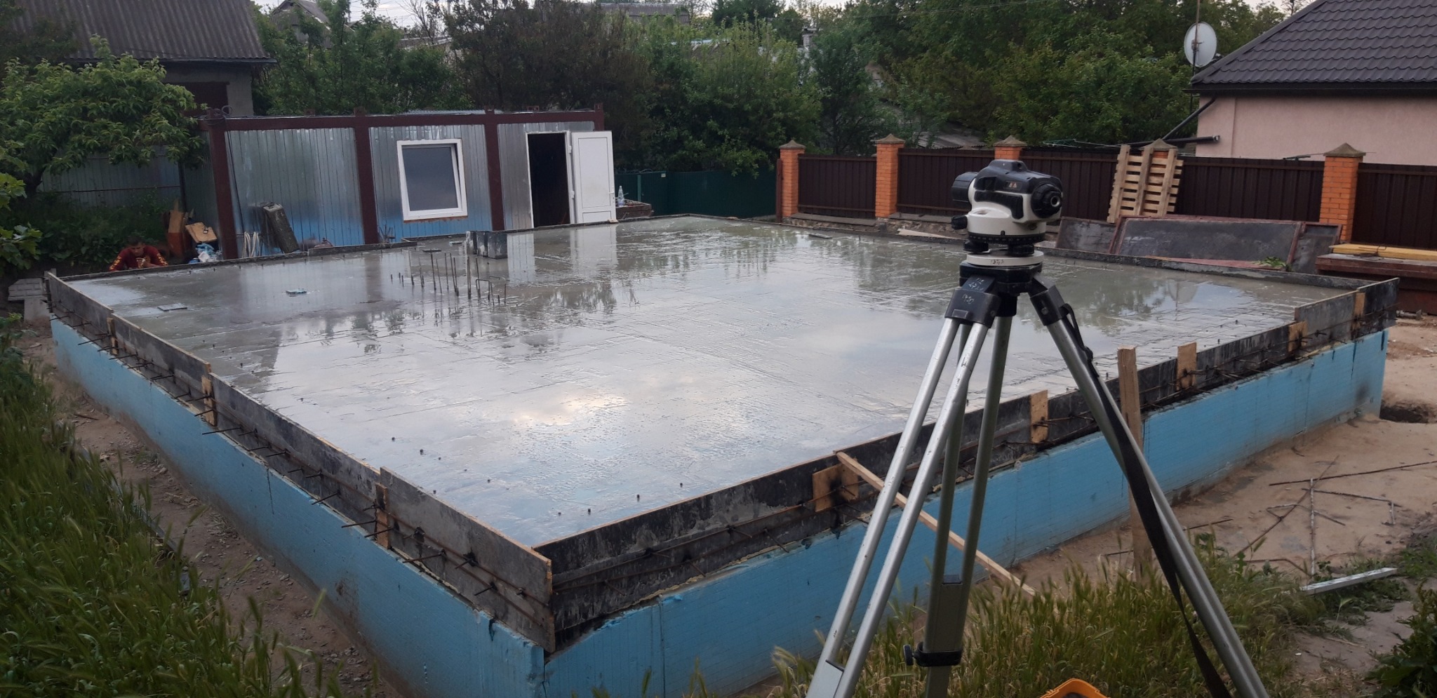 Świeżo wylana, mokra posadzka zbrojona, otoczona szalunkiem z niebieską izolacją, widoczny niwelator optyczny na statywie, w tle budynek gospodarczy i ogrodzenie z cegły.
