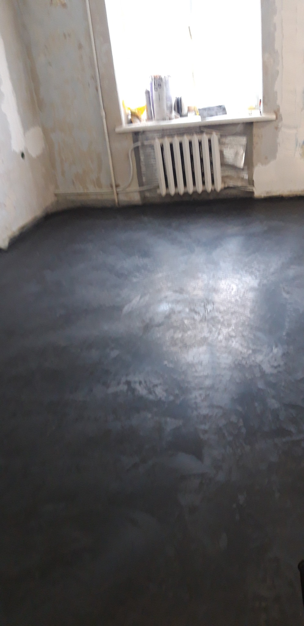 Świeżo wylana, ciemnoszara posadzka żywiczna w surowym wnętrzu z grzejnikiem pod oknem, widoczne niedoskonałości na ścianach.