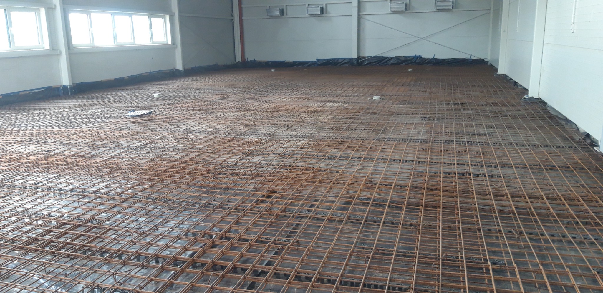 Przygotowanie podłoża pod posadzkę w hali przemysłowej, widoczna siatka zbrojeniowa na folii izolacyjnej, w tle białe ściany i okna.