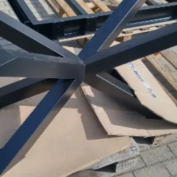 Czarna, metalowa podstawa stołu o geometrycznym kształcie gwiazdy, sfotografowana na zewnątrz na palecie i kartonie, z widocznymi spawami i mocowaniami.
