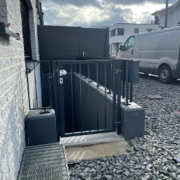 Zabezpieczenie zejścia do piwnicy z czarną metalową kratą i furtką, widok z poziomu gruntu, obok budynku z białej cegły, na pierwszym planie metalowy stolik z dwoma szklankami.