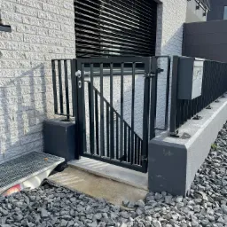 Nowoczesna, czarna furtka z pionowych profili prowadząca do zejścia do piwnicy, zamontowana przy jasnym, ceglanym budynku, otoczona szarym kamieniem.