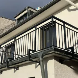 Czarna, metalowa balustrada balkonowa z pionowymi prętami, zamocowana na wspornikach do ściany budynku z orynnowaniem.