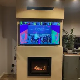 Nowoczesny kominek z czarną ramą w zabudowie z gładzi, nad nim telewizor z transmisją sportową i oświetlenie punktowe na suficie.