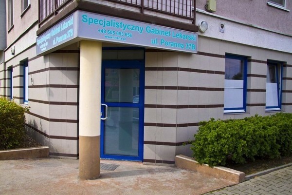Fasada budynku z szyldem 'Specjalistyczny Gabinet Lekarski ul. Poranna 37B' nad niebieskimi drzwiami wejściowymi, otoczona zielenią.