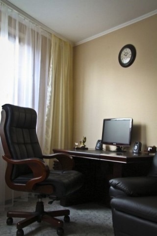 Gabinet: czarne skórzane krzesło obrotowe, biurko z komputerem i głośnikami, zegar na ścianie, neutralne kolory ścian i zasłon.