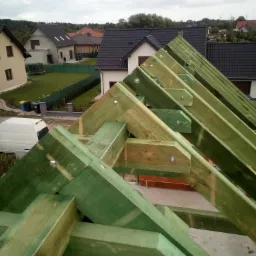 Zielona, impregnowana konstrukcja drewniana dachu z widocznymi śrubami mocującymi, na tle domów jednorodzinnych i pochmurnego nieba.