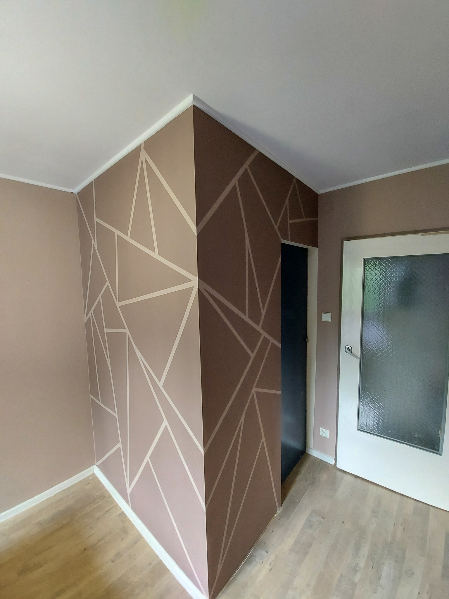 Ściana w pokoju pomalowana w geometryczne wzory w odcieniach beżu i brązu, z widocznym fragmentem podłogi z paneli i drzwiami.