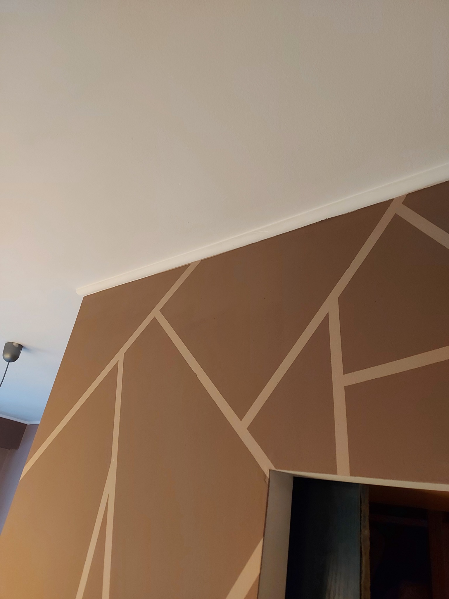 Brązowa ściana z geometrycznym wzorem z jasnych pasków, biały sufit z listwą przypodłogową, widoczny fragment lampy i futryny drzwi.