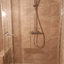 Kabina prysznicowa z transparentnego szkła z chromowaną armaturą na tle beżowych płytek imitujących marmur.