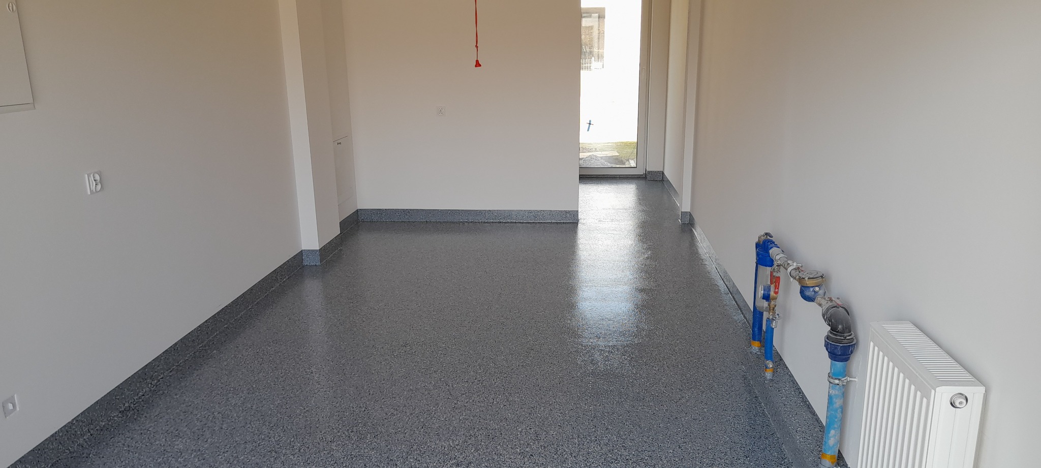 Wnętrze pustego pomieszczenia z szarą, żywiczną posadzką z drobnymi plamkami, widoczne rury i grzejnik przy ścianie, świeżo wykończone.
