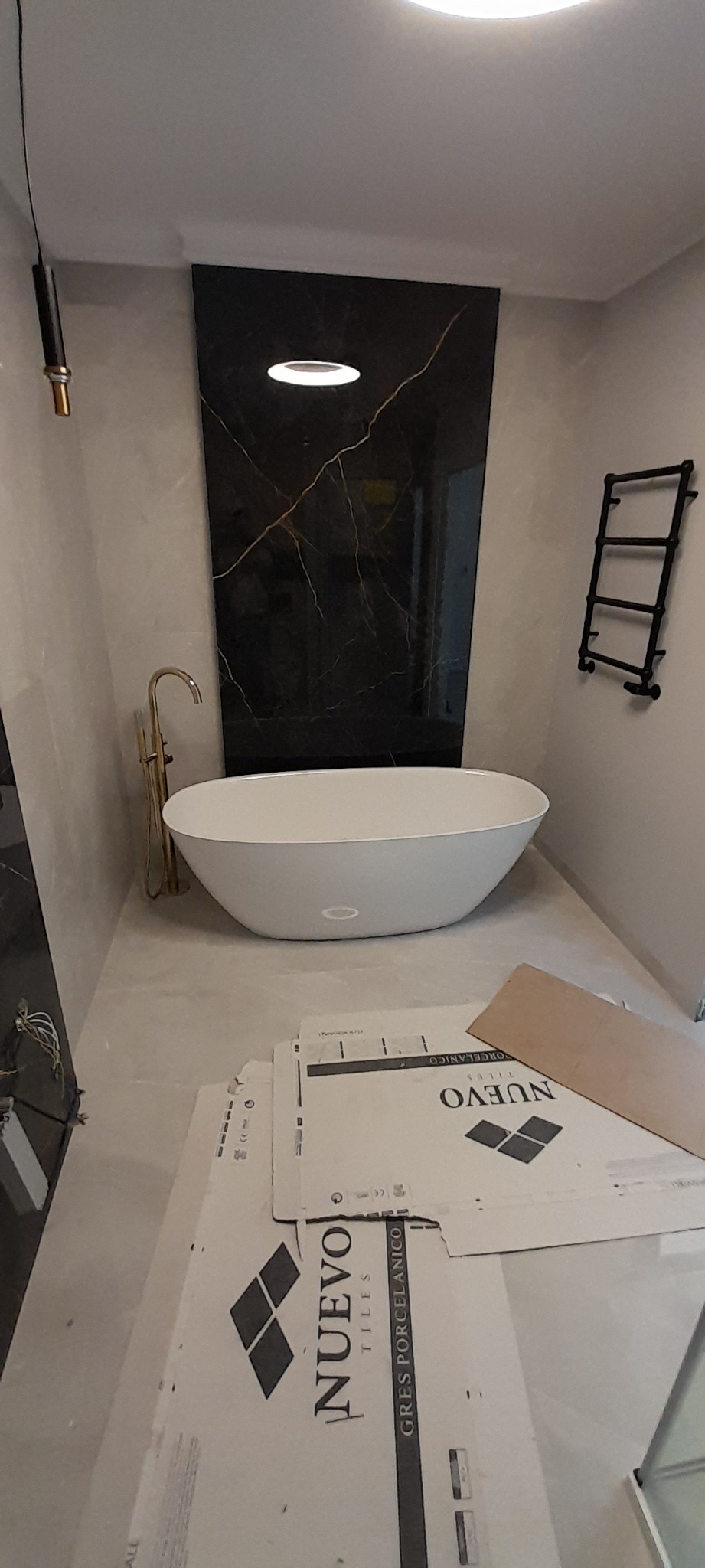 Nowoczesna łazienka w trakcie wykończenia: wolnostojąca wanna, złota bateria, czarny panel z marmurowym wzorem, grzejnik drabinkowy, kartony z płytkami Nuevo Tiles na podłodze.