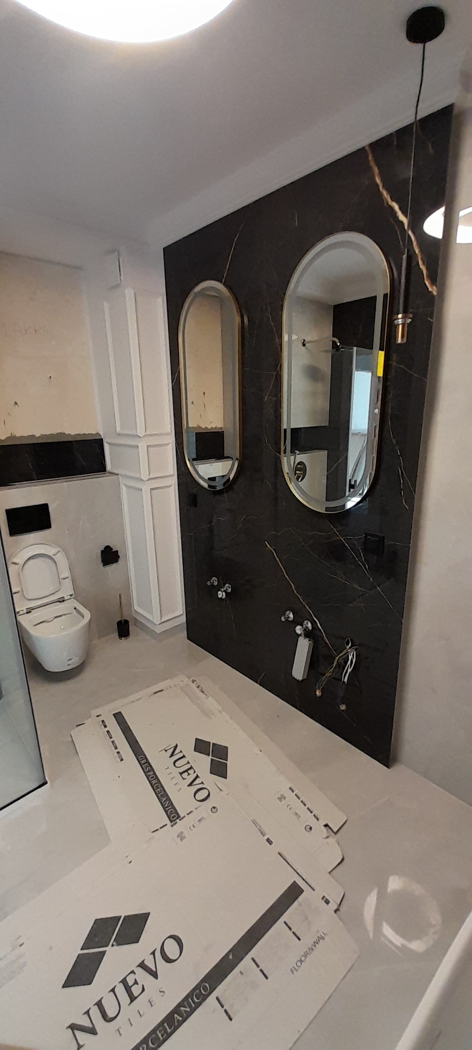 Łazienka w trakcie aranżacji: czarna ściana z marmurowym wzorem, owalne lustra z podświetleniem, wisząca toaleta, białe panele ścienne i kartony płytek Nuevo Tiles na podłodze.