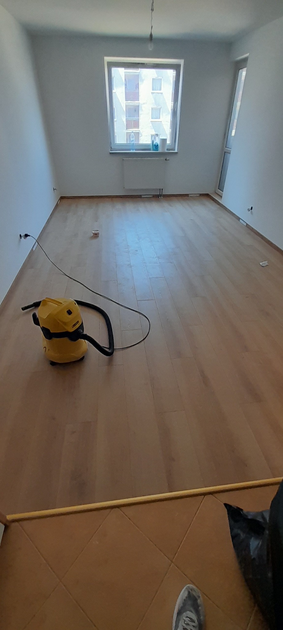 Puste, jasne pomieszczenie z drewnianą podłogą, częściowo odkurzone żółtym odkurzaczem przemysłowym; widoczne białe ściany, okno z widokiem na budynek mieszkalny i fragment brązowych płytek...