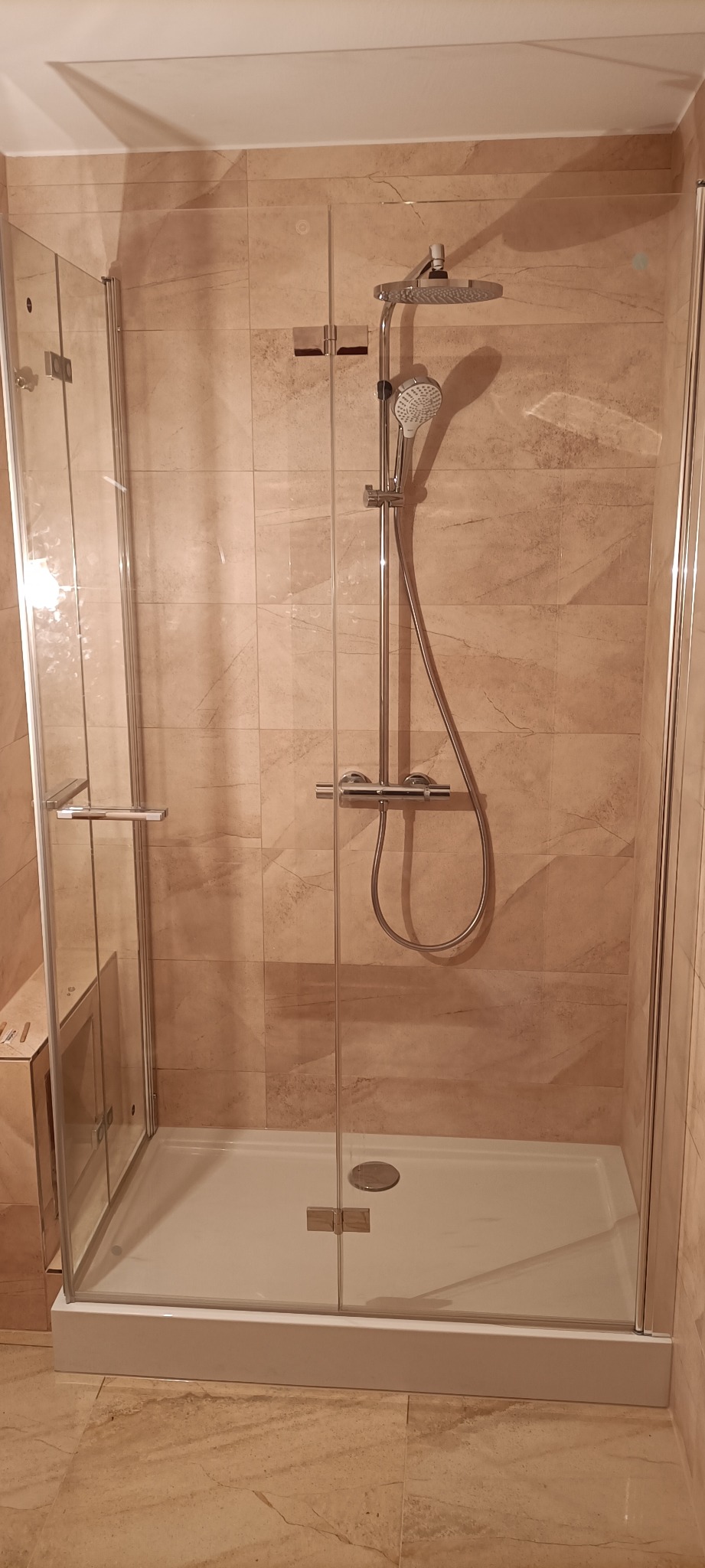 Kabina prysznicowa z transparentnego szkła z chromowaną armaturą na tle beżowych płytek imitujących marmur.