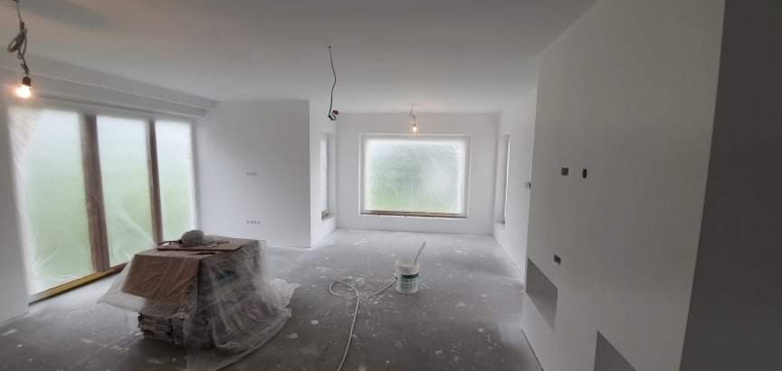 Wnętrze w trakcie malowania: okna zabezpieczone folią, wiadro z farbą na podłodze, zakurzone podłoże, widoczne kable oświetleniowe zwisające z sufitu.