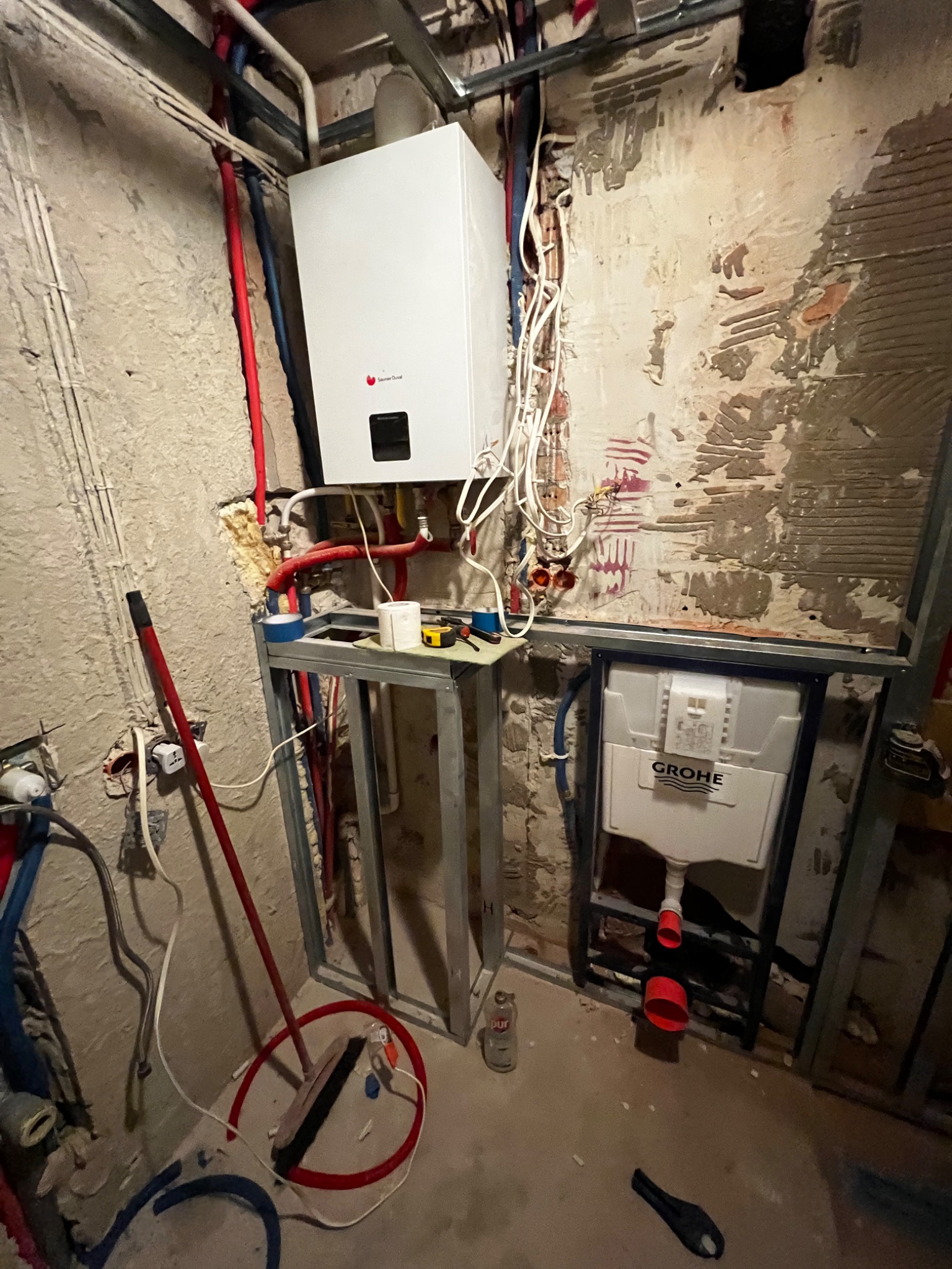 Montaż instalacji wodno-kanalizacyjnej w surowym stanie, widoczny stelaż podtynkowy Grohe, wiszący bojler Saunier Duval, rury w kolorze czerwonym i niebieskim, ściany w trakcie przygotowania...