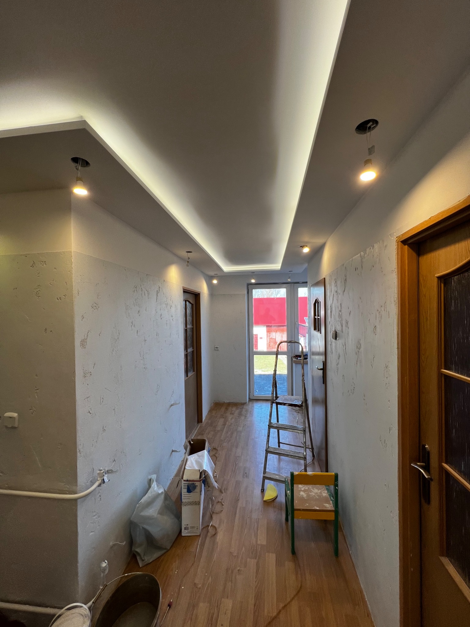 Korytarz w trakcie prac wykończeniowych: szpachlowane ściany, podwieszony sufit z oświetleniem LED, drabina, wiadro z narzędziami i materiałami budowlanymi na podłodze.
