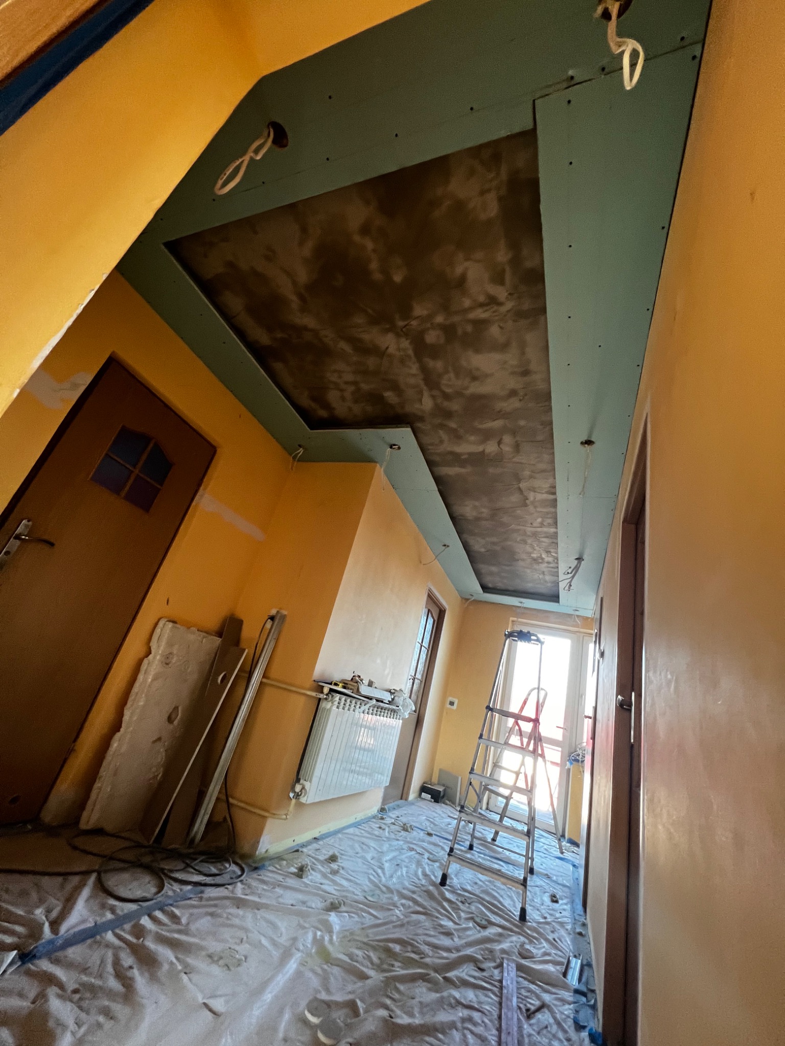 Wnętrze w trakcie montażu podwieszanego sufitu z płyt gipsowo-kartonowych na metalowej konstrukcji, zabezpieczone folią malarską, z widoczną drabiną i przewodami elektrycznymi.
